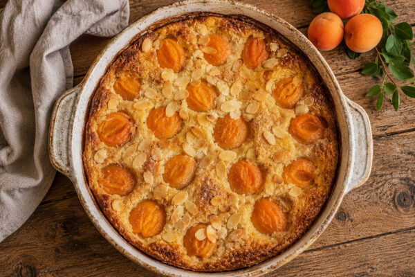 Draufsicht auf den fertig gebackenen Aprikosen-Mandel-Clafoutis in derselben hellen runden Keramikform mit goldbrauner Oberfläche und Mandelblättchen.
