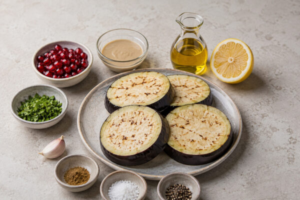 Zutaten- oder Vorbereitungsansicht mit Auberginen, Tahini, Zitronensaft, Olivenöl, Knoblauch, Granatapfelkernen, Petersilie, Kreuzkümmel, Salz und Pfeffer.