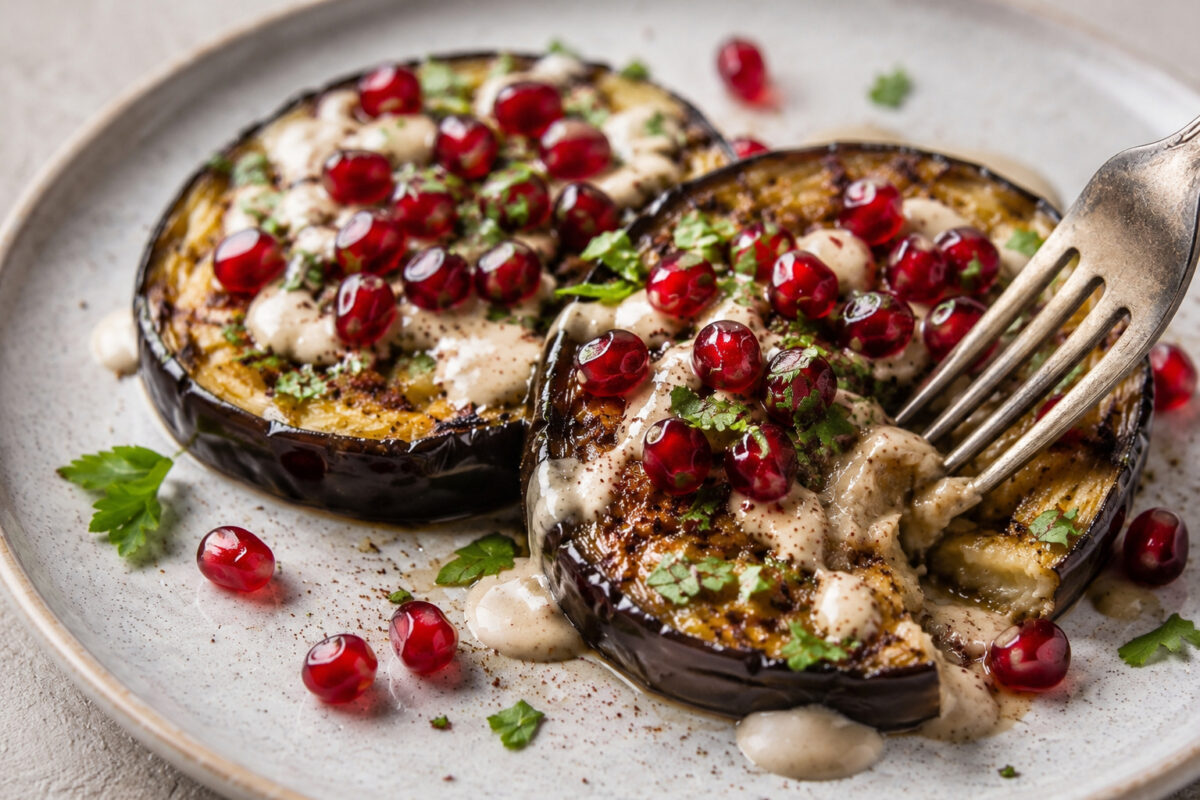Nahaufnahme beziehungsweise Servierszene der Auberginensteaks mit Tahini-Sauce und Granatapfelkernen, mit einer Gabel als Serviermoment.