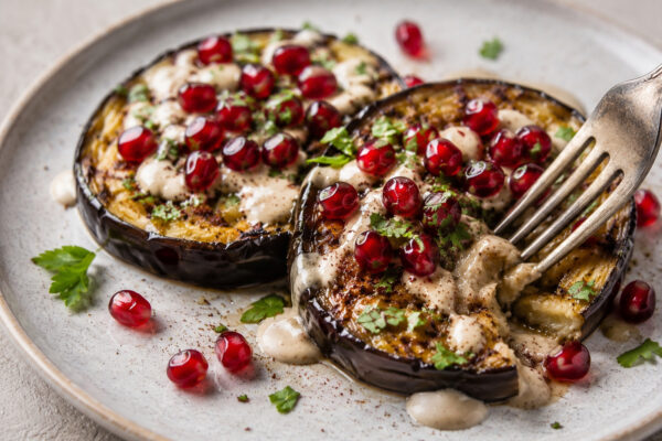 Nahaufnahme beziehungsweise Servierszene der Auberginensteaks mit Tahini-Sauce und Granatapfelkernen, mit einer Gabel als Serviermoment.