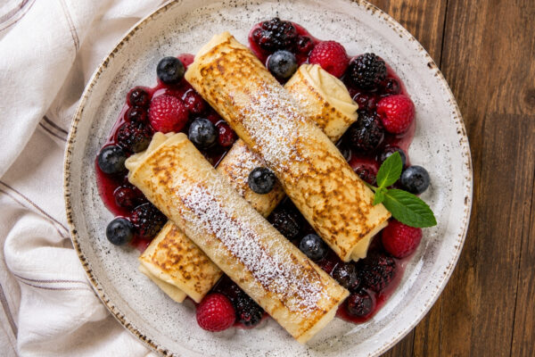 Draufsicht auf mehrere goldgelbe Blintzes mit Quarkfüllung und Beerenkompott auf einem hellen Teller.