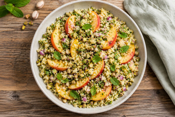 Draufsicht auf das fertig angerichtete Gericht in derselben großen hellen Keramikschale: lockerer Couscous-Salat mit Pfirsichspalten, Pistazien, roten Zwiebelwürfeln und frischen Kräutern. Rustikaler Holztisch, hellgrünes Tuch rechts, Minze und Pistazien…