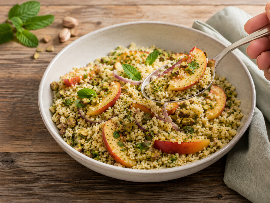 Nahe Servieransicht des fertigen Gerichts in derselben großen hellen Keramikschale: Ein Löffel hebt eine Portion Couscous-Salat mit Pfirsichspalte, Pistazien und Kräutern an. Das hellgrüne Tuch rechts und Minze mit Pistazien links oben bleiben sichtbar.
