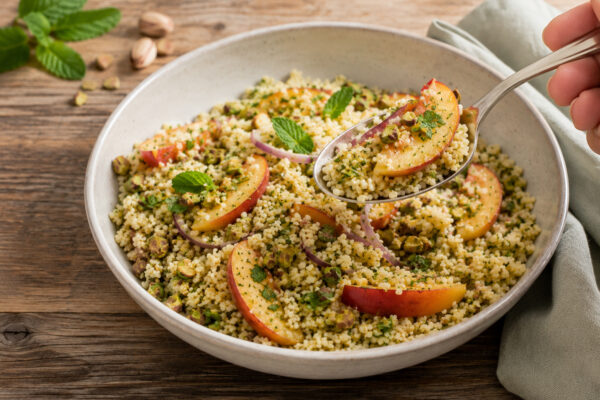 Nahe Servieransicht des fertigen Gerichts in derselben großen hellen Keramikschale: Ein Löffel hebt eine Portion Couscous-Salat mit Pfirsichspalte, Pistazien und Kräutern an. Das hellgrüne Tuch rechts und Minze mit Pistazien links oben bleiben sichtbar.