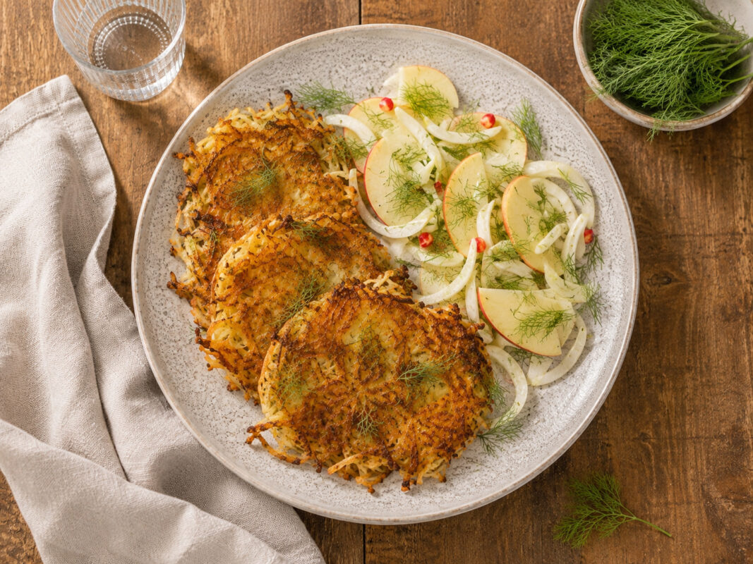 Draufsicht auf das fertig angerichtete Gericht auf demselben großen hellen Teller: mehrere goldbraune Fenchel-Kartoffel-Rösti mit einem frischen Apfel-Fenchel-Dill-Salat. Rustikaler Holztisch, Leinentuch links, Wasserglas hinten links und kleine Dillscha…