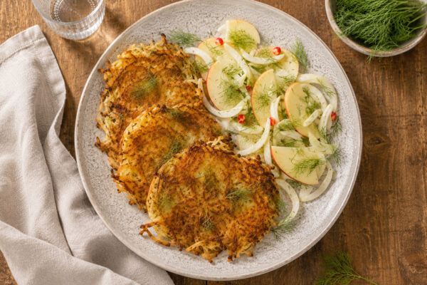 Draufsicht auf das fertig angerichtete Gericht auf demselben großen hellen Teller: mehrere goldbraune Fenchel-Kartoffel-Rösti mit einem frischen Apfel-Fenchel-Dill-Salat. Rustikaler Holztisch, Leinentuch links, Wasserglas hinten links und kleine Dillscha…