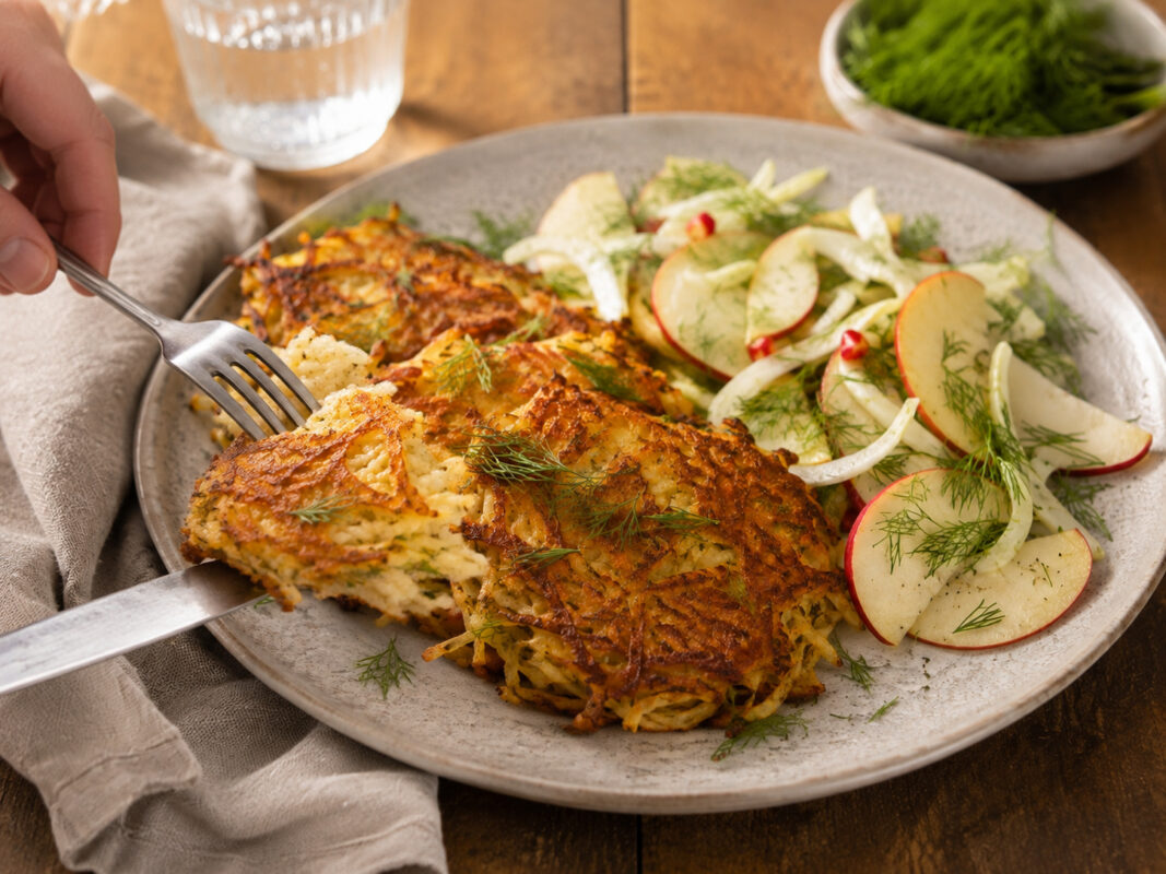 Serviersituation des fertigen Gerichts auf demselben großen hellen Teller: eine Portion Rösti wird mit neutralem Besteck angehoben oder angeschnitten, dazu liegt der frische Apfel-Fenchel-Dill-Salat daneben. Die warme natürliche Foodfotografie-Anmutung u…