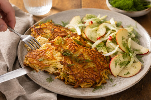 Serviersituation des fertigen Gerichts auf demselben großen hellen Teller: eine Portion Rösti wird mit neutralem Besteck angehoben oder angeschnitten, dazu liegt der frische Apfel-Fenchel-Dill-Salat daneben. Die warme natürliche Foodfotografie-Anmutung u…