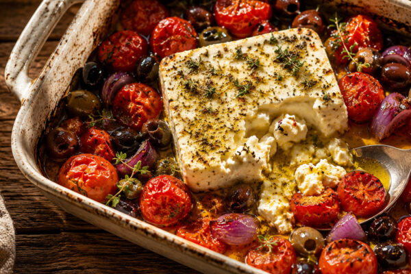 Fertiger gebackener Feta mit aufgeplatzten Tomaten, Oliven und Zwiebel; Servieransicht mit entnommener Portion.