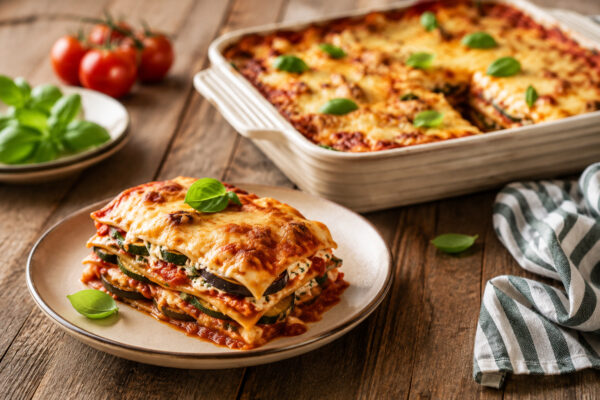 Fertige Gemüse-Lasagne als angerichtete Portion auf einem Teller; sichtbare Schichten aus Pasta, Aubergine, Zucchini, Tomatensauce und Käse, mit der restlichen Lasagne in der Auflaufform im Hintergrund.