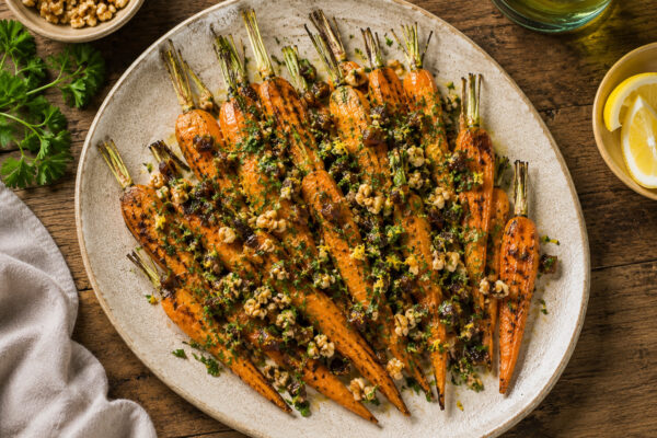 Draufsicht auf das fertig angerichtete Gericht auf derselben großen ovalen hellen Platte: geröstete Möhren mit Dattel-Walnuss-Gremolata, Petersilie und Zitronenzesten, in gleicher warmer natürlicher Foodfotografie und mit denselben Requisiten.