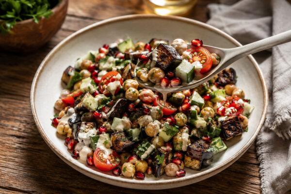 Serviersituation mit einer Löffelportion aus derselben hellen Keramikschale; der Löffel zeigt geröstete Auberginenwürfel, Kichererbsen, Gurke, Kirschtomaten, Granatapfel und Tahini-Dressing.