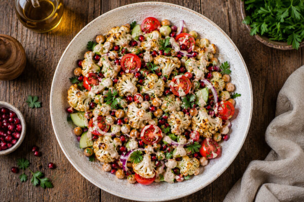 Fertiger gerösteter Blumenkohlsalat in derselben großen hellen gesprenkelten Keramikschale in Draufsicht, mit vollständig sichtbarer Schale, geröstetem Blumenkohl, Kichererbsen, Gurke, Kirschtomaten, roten Zwiebeln, Granatapfel und hellem Tahini-Dressing.