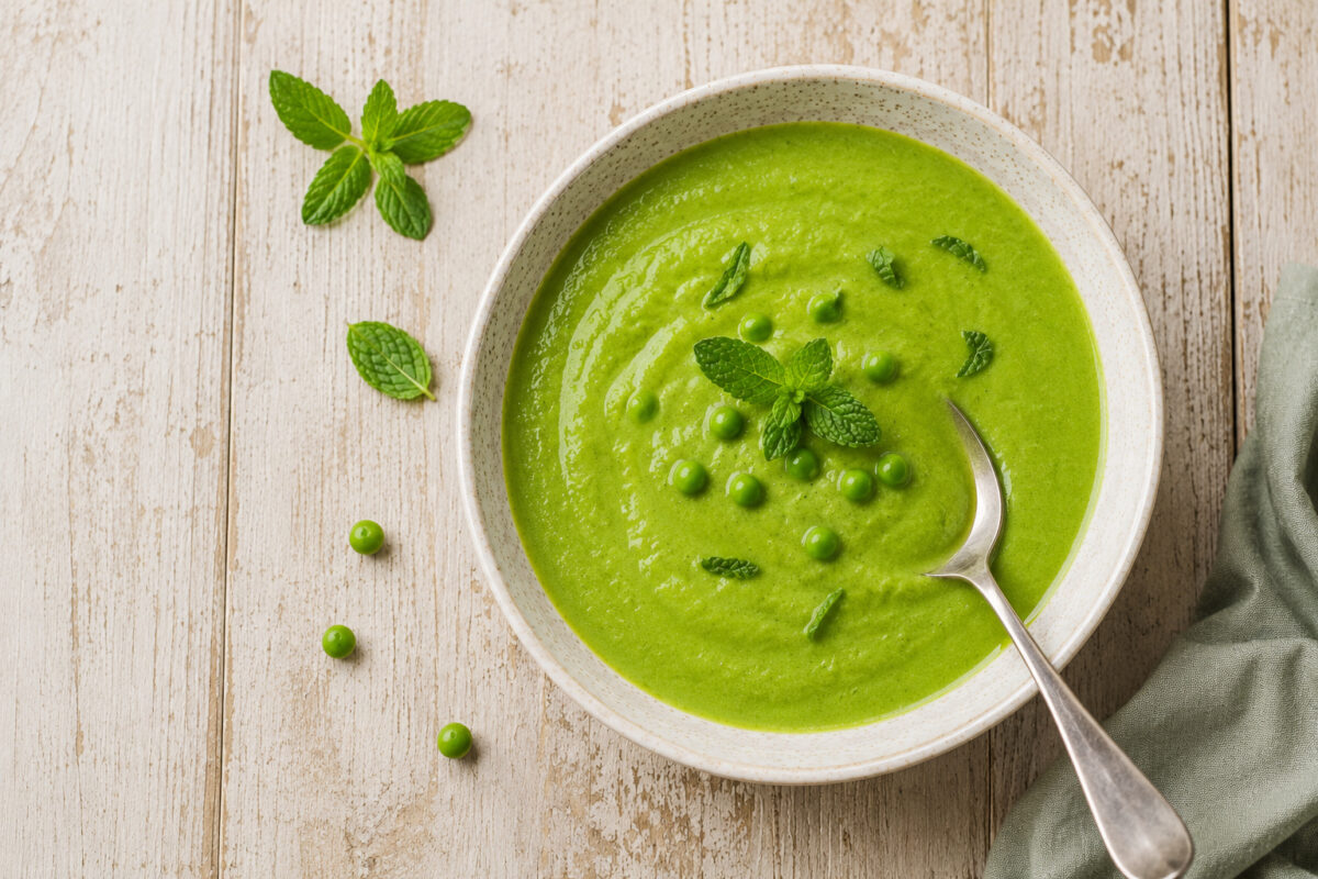 Fertige Erbsensuppe mit Minze aus deutlich stärkerer Aufsicht, mit Löffel und gleicher Keramik-, Licht- und Farbwelt.