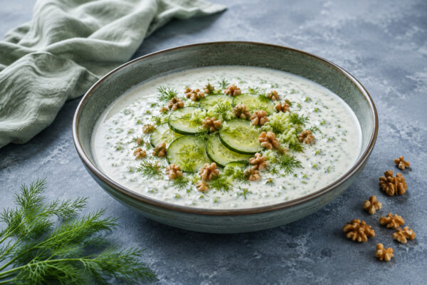 Fertig angerichtete Gurken-Joghurt-Suppe mit Gurkenscheiben, Dill, geriebener Gurke und Walnüssen in einer flachen Schale.