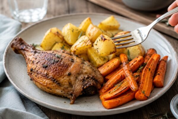 Lebendige Servierszene für „Hähnchenkeule mit Kartoffeln und Ofenmöhren“: eine Gabel oder ein Besteckteil nimmt gerade eine Portion auf; Hähnchenkeule, Kartoffeln und Möhren bleiben trotzdem klar erkennbar.