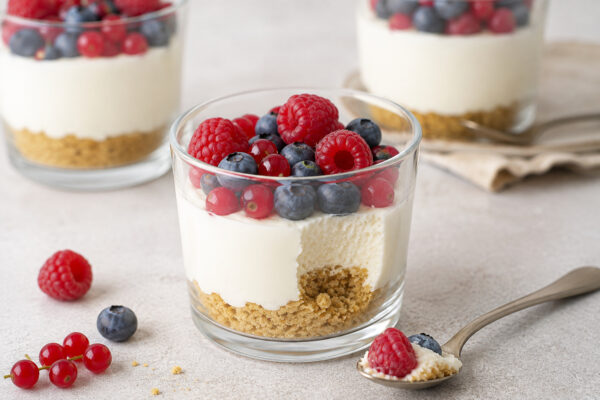 Servierszene im Detail: Käsekuchen im Glas mit sichtbarer Schichtstruktur, Beerentopping und Dessertlöffel mit entnommener Portion.