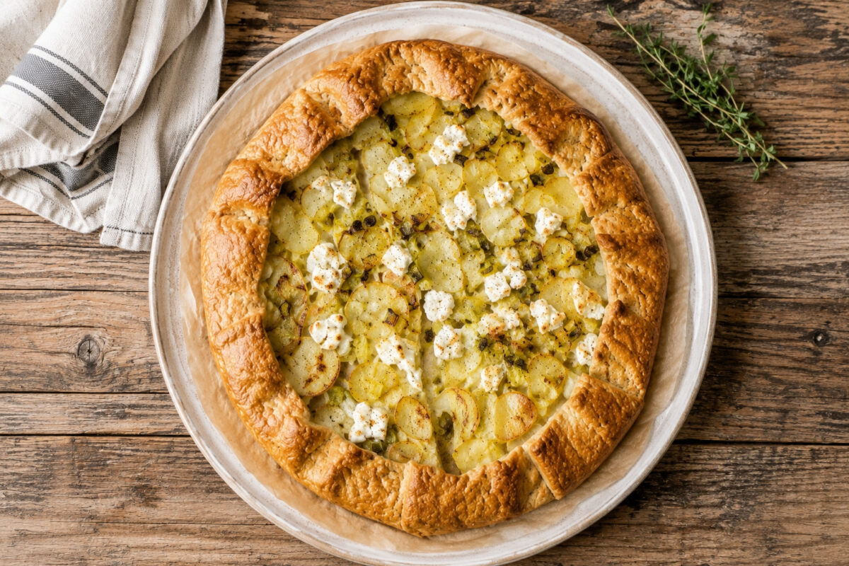 Draufsicht auf die fertig gebackene Kartoffel-Lauch-Galette mit Ziegenkäse in derselben hellen runden Platte; die gesamte Galette ist vollständig sichtbar, mit goldbraunem Teigrand, Kartoffelscheiben, Lauch und Ziegenkäse.