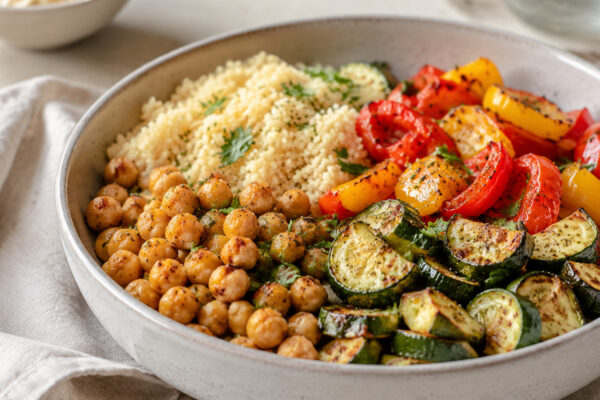 Alternative Perspektive des fertigen Gerichts: Echte Bowl mit klar getrennten Segmenten aus Kichererbsen, Couscous und Paprika und Zucchini; Zutaten erst beim Essen mischbar und optisch deutlich voneinander abgegrenzt. Etwas näher aufgenommen, natürliche…