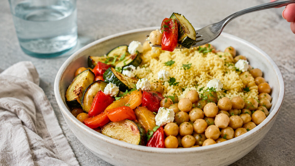 Lebendige Servierszene für „Kichererbsen-Couscous-Bowl mit Ofengemüse“: eine Gabel oder ein Besteckteil nimmt gerade eine Portion auf; Kichererbsen, Couscous und Paprika und Zucchini bleiben trotzdem klar erkennbar.