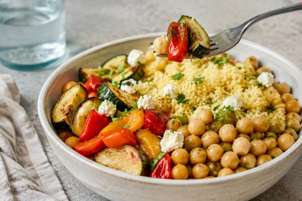 Lebendige Servierszene für „Kichererbsen-Couscous-Bowl mit Ofengemüse“: eine Gabel oder ein Besteckteil nimmt gerade eine Portion auf; Kichererbsen, Couscous und Paprika und Zucchini bleiben trotzdem klar erkennbar.
