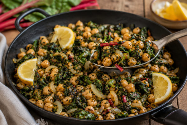 Nahe Servieransicht des fertigen Gerichts in derselben großen schwarzen Pfanne: eine Portion aus Kichererbsen, Mangold und Zwiebeln wird mit neutralem Metalllöffel angehoben; Zitronenspalten und die warme natürliche Foodfotografie-Anmutung bleiben erhalt…