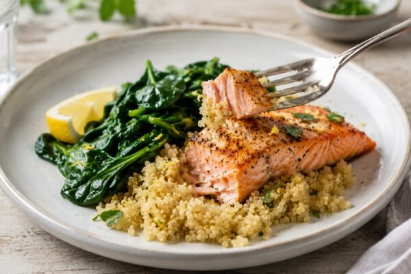 Lebendige Servierszene für „Lachs auf Zitronenquinoa mit Blattspinat“: eine Gabel oder ein Besteckteil nimmt gerade eine Portion auf; Lachsfilet, Quinoa und Blattspinat bleiben trotzdem klar erkennbar.