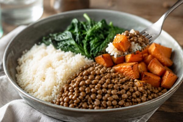 Lebendige Servierszene für „Linsen-Reis-Bowl mit Spinat und Möhren“: eine Gabel oder ein Besteckteil nimmt gerade eine Portion auf; braune Linsen, Reis und Blattspinat und Möhren bleiben trotzdem klar erkennbar.