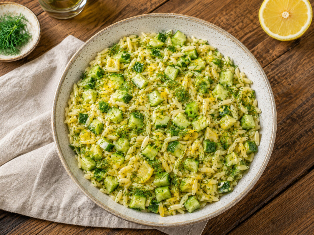 Draufsicht auf den fertig angerichteten Orzo-Salat mit Gurke, Zitrone und Dill in derselben großen hellen gesprenkelten Keramikschale. Die Schale ist vollständig sichtbar, der Salat wirkt frisch und locker, mit Gurkenwürfeln, Zitronenstückchen und reichl…