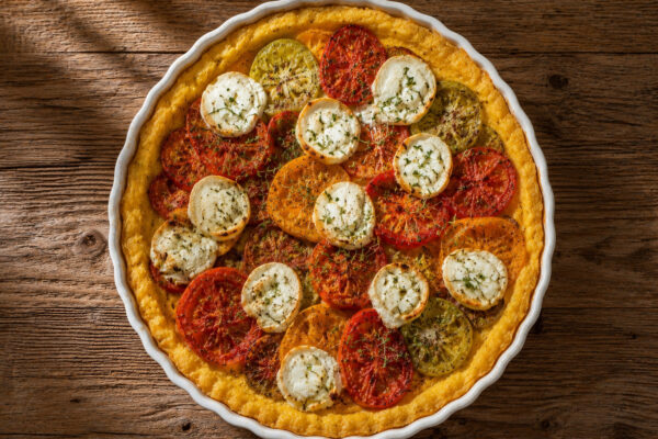 Draufsicht auf die fertig gebackene Polenta-Tarte in derselben weißen geriffelten Tarteform mit bunten Tomaten, Ziegenkäse und Thymian.
