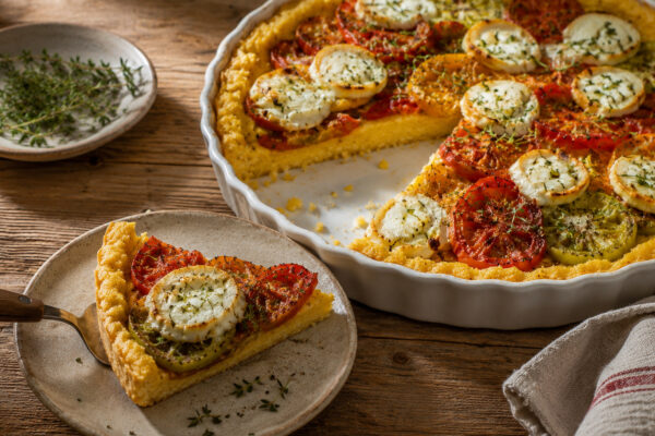 Serviersituation mit einem Stück Polenta-Tarte auf einem hellen Teller; die angeschnittene Tarteform mit Tomaten, Ziegenkäse und Thymian steht im Hintergrund.