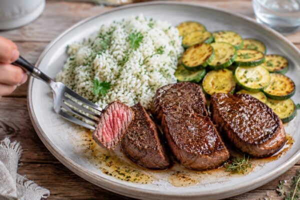 Lebendige Servierszene für „Rindermedaillons mit Kräuterreis und Zucchini“: eine Gabel oder ein Besteckteil nimmt gerade eine Portion auf; Rindermedaillons, Reis und Zucchini bleiben trotzdem klar erkennbar.
