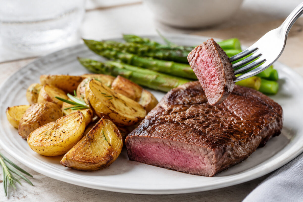 Lebendige Servierszene für „Rindersteak mit Rosmarinkartoffeln und grünem Spargel“: eine Gabel oder ein Besteckteil nimmt gerade eine Portion auf; Rindersteak, Kartoffeln und grüner Spargel bleiben trotzdem klar erkennbar.