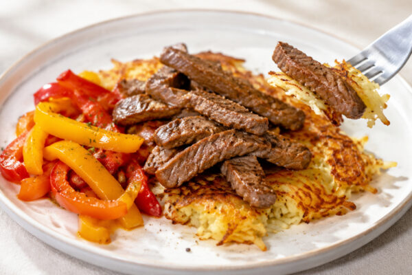 Lebendige Servierszene für „Rinderstreifen auf Kartoffelrösti mit Paprikagemüse“: eine Gabel oder ein Besteckteil nimmt gerade eine Portion auf; Rinderstreifen, Kartoffeln und Paprika bleiben trotzdem klar erkennbar.