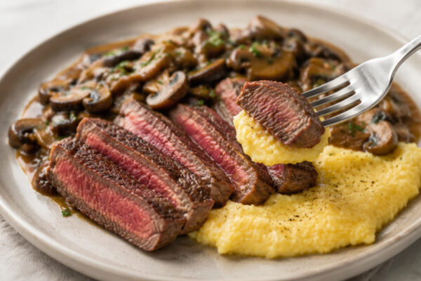 Lebendige Servierszene für „Rindfleisch auf Polenta mit Pilzragout“: eine Gabel oder ein Besteckteil nimmt gerade eine Portion auf; Rinderhüftsteak, Polenta und Champignons bleiben trotzdem klar erkennbar.