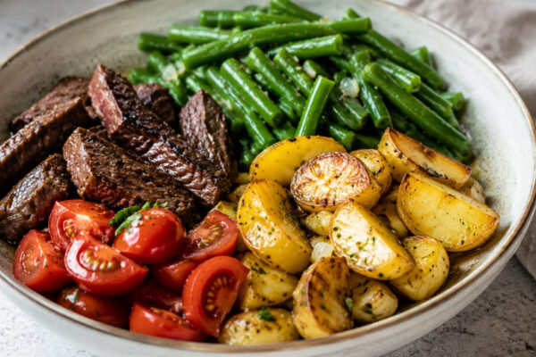 Alternative Perspektive des fertigen Gerichts: Echte Bowl mit klar getrennten Segmenten aus Rinderstreifen, Kartoffeln und grüne Bohnen und Tomaten; Zutaten erst beim Essen mischbar und optisch deutlich voneinander abgegrenzt. Etwas näher aufgenommen, na…