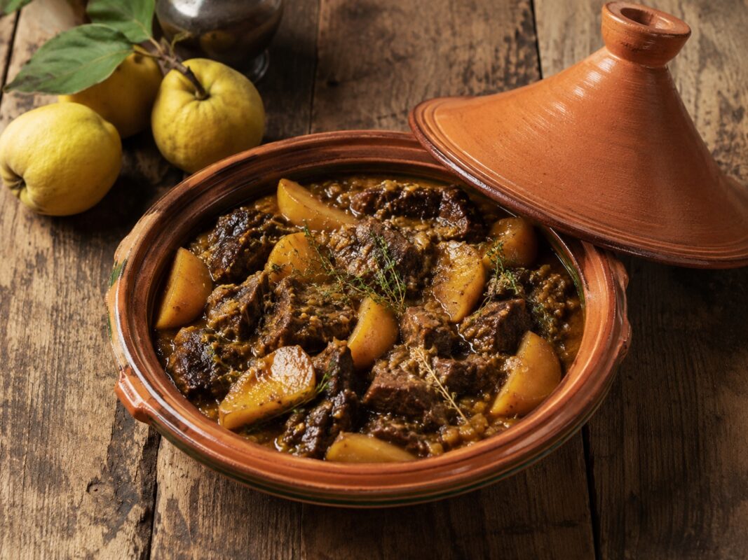 Terrakotta-Tajine mit dunklem Rinderschmorgericht, goldenen Quittenspalten und Kräutern auf rustikalem Holztisch.