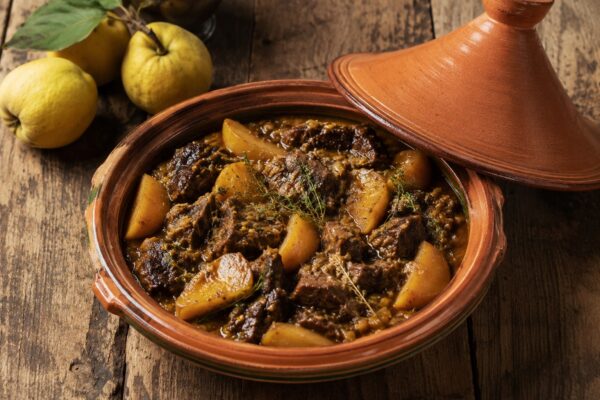 Terrakotta-Tajine mit dunklem Rinderschmorgericht, goldenen Quittenspalten und Kräutern auf rustikalem Holztisch.