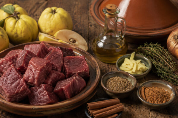 Zutaten- und Vorbereitungsansicht mit Rindfleischwürfeln, Quitten, Zwiebeln, Olivenöl, Brühe, Zimt, Kreuzkümmel, Ingwer und Kräutern neben der Terrakotta-Tajine.