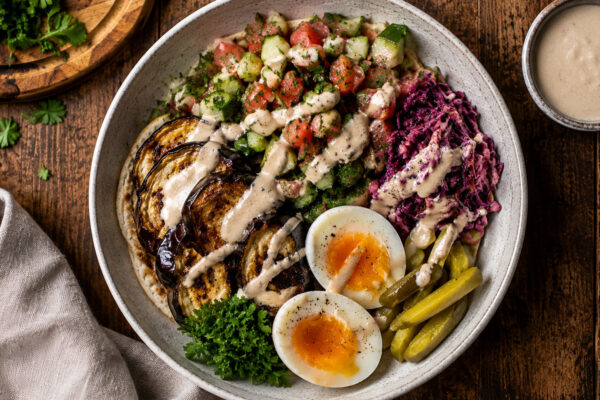 Draufsicht auf die fertige Sabich-Schale mit gerösteter Aubergine, Eiern, Gurken-Tomaten-Salat, Rotkohlsalat, Essiggurken und Tahini-Sauce.