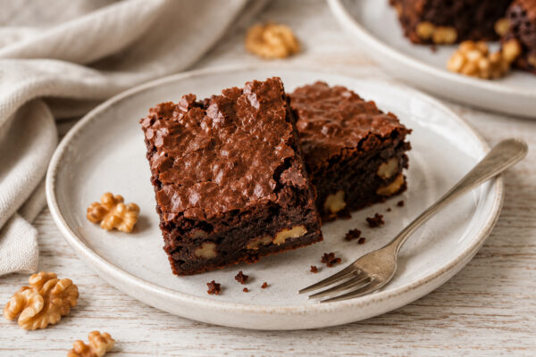 Servierszene mit zwei fertigen Brownies, sichtbarer fudgiger Innenstruktur, Walnüssen, Krümeln und Gabel auf hellem Keramikteller.