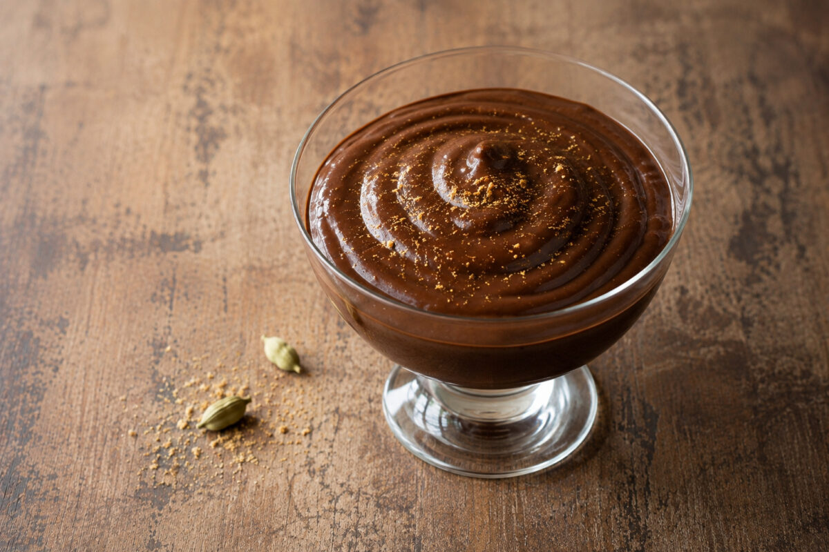 Fertiger Schokoladenpudding mit Kardamom in derselben Glasschale auf rustikalem Holz, aus leicht anderer Perspektive mit Kardamomkapseln als Requisite.