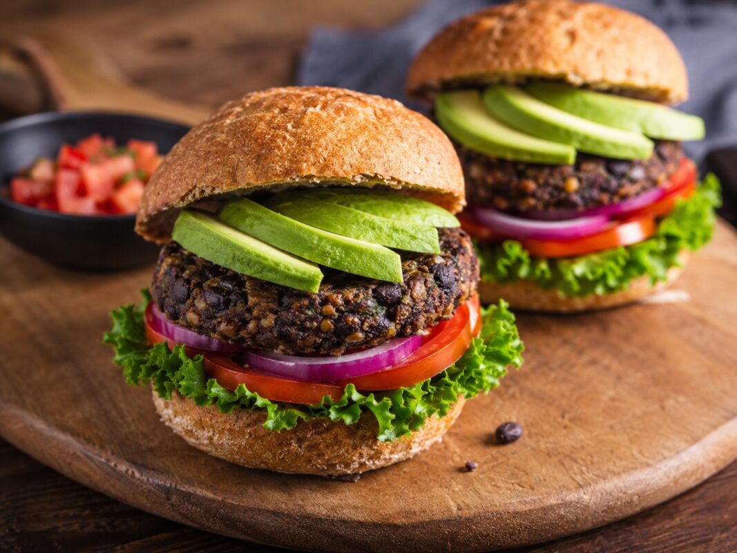 Zwei schwarze-Bohnen-Burger auf Holzbrett mit Avocadoscheiben, Tomate, Zwiebeln und Salat.