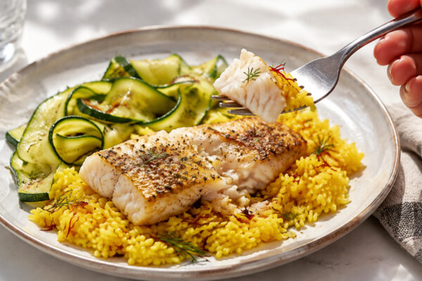 Lebendige Servierszene für „Seelachs auf Safranreis mit Zucchinibändern“: eine Gabel oder ein Besteckteil nimmt gerade eine Portion auf; Seelachsfilet, Reis und Zucchini bleiben trotzdem klar erkennbar.