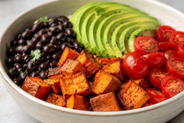 Alternative Perspektive des fertigen Gerichts: Echte Bowl mit klar getrennten Segmenten aus schwarze Bohnen, Süßkartoffeln und Avocado und Tomaten; Zutaten erst beim Essen mischbar und optisch deutlich voneinander abgegrenzt. Etwas näher aufgenommen, nat…