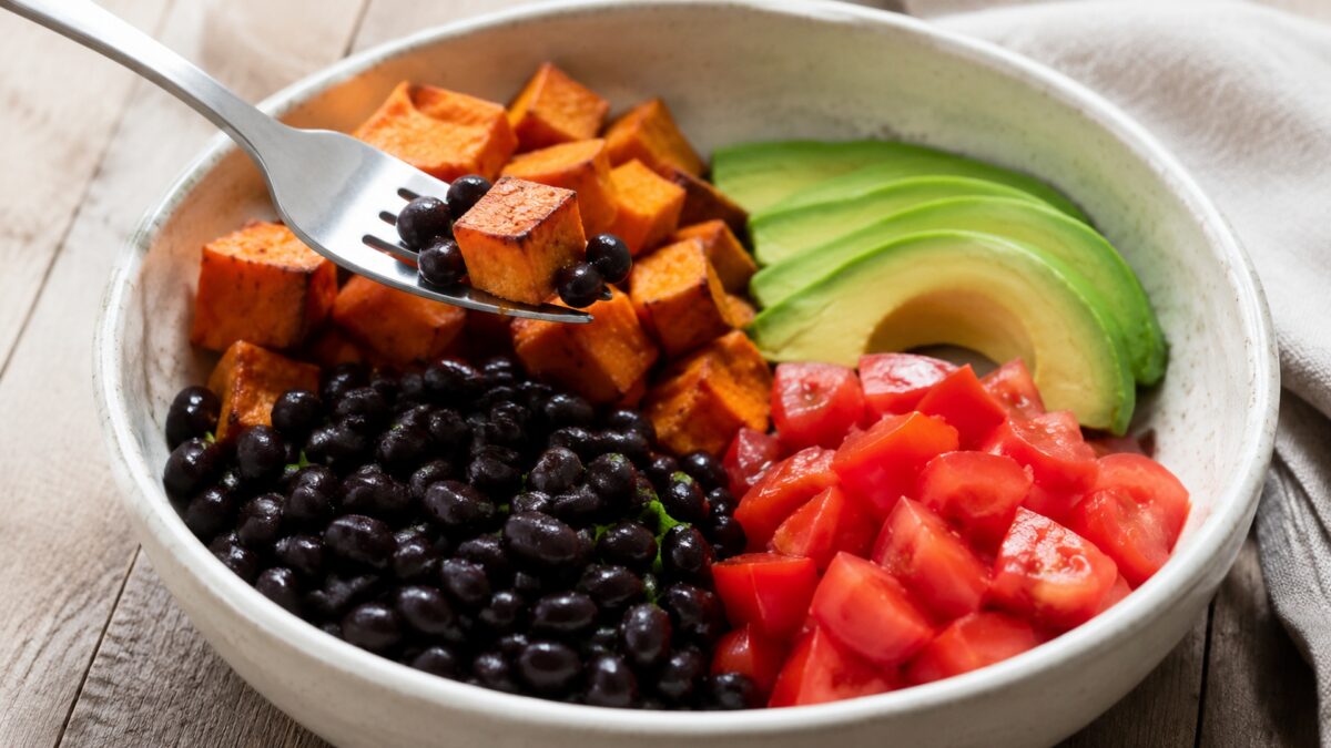 Lebendige Servierszene für „Süßkartoffel-Bohnen-Bowl mit Avocado“: eine Gabel oder ein Besteckteil nimmt gerade eine Portion auf; schwarze Bohnen, Süßkartoffeln und Avocado und Tomaten bleiben trotzdem klar erkennbar.