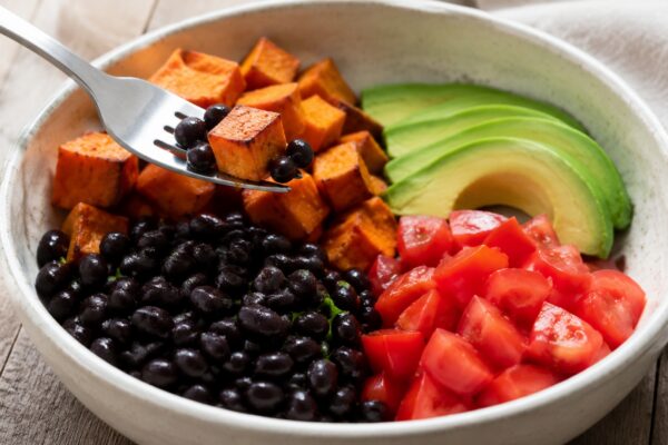 Lebendige Servierszene für „Süßkartoffel-Bohnen-Bowl mit Avocado“: eine Gabel oder ein Besteckteil nimmt gerade eine Portion auf; schwarze Bohnen, Süßkartoffeln und Avocado und Tomaten bleiben trotzdem klar erkennbar.