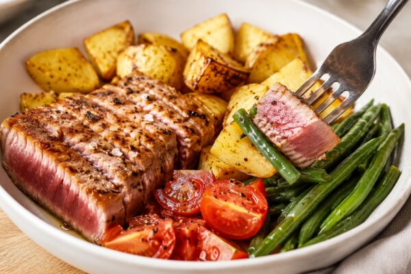 Lebendige Servierszene für „Thunfisch-Kartoffel-Bowl mit grünen Bohnen“: eine Gabel oder ein Besteckteil nimmt gerade eine Portion auf; Thunfischsteak, Kartoffeln und grüne Bohnen und Tomaten bleiben trotzdem klar erkennbar.