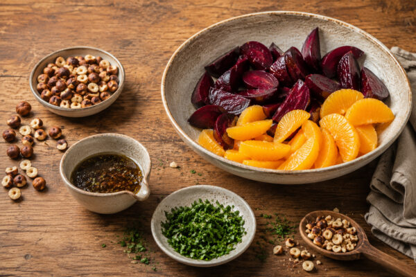Zutaten-/Vorbereitungsansicht mit Roter Bete, Orangenfilets, gerösteten Haselnüssen, gehackter Petersilie und Dressing in derselben rustikalen Material- und Lichtwelt.