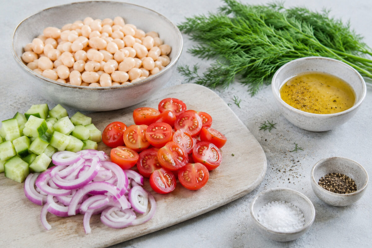 Zutaten- und Vorbereitungsansicht mit weißen Bohnen, halbierten Tomaten, Gurke, roten Zwiebeln, Dill und Dressing auf hellem Steinuntergrund.